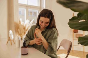 cheerful-young-caucasian-woman-applies-cream-her-hands-while-sitting-table-light-room-brunette-girl-with-smooth-skin-wears-shirt-wellness-self-care-concept_11zon